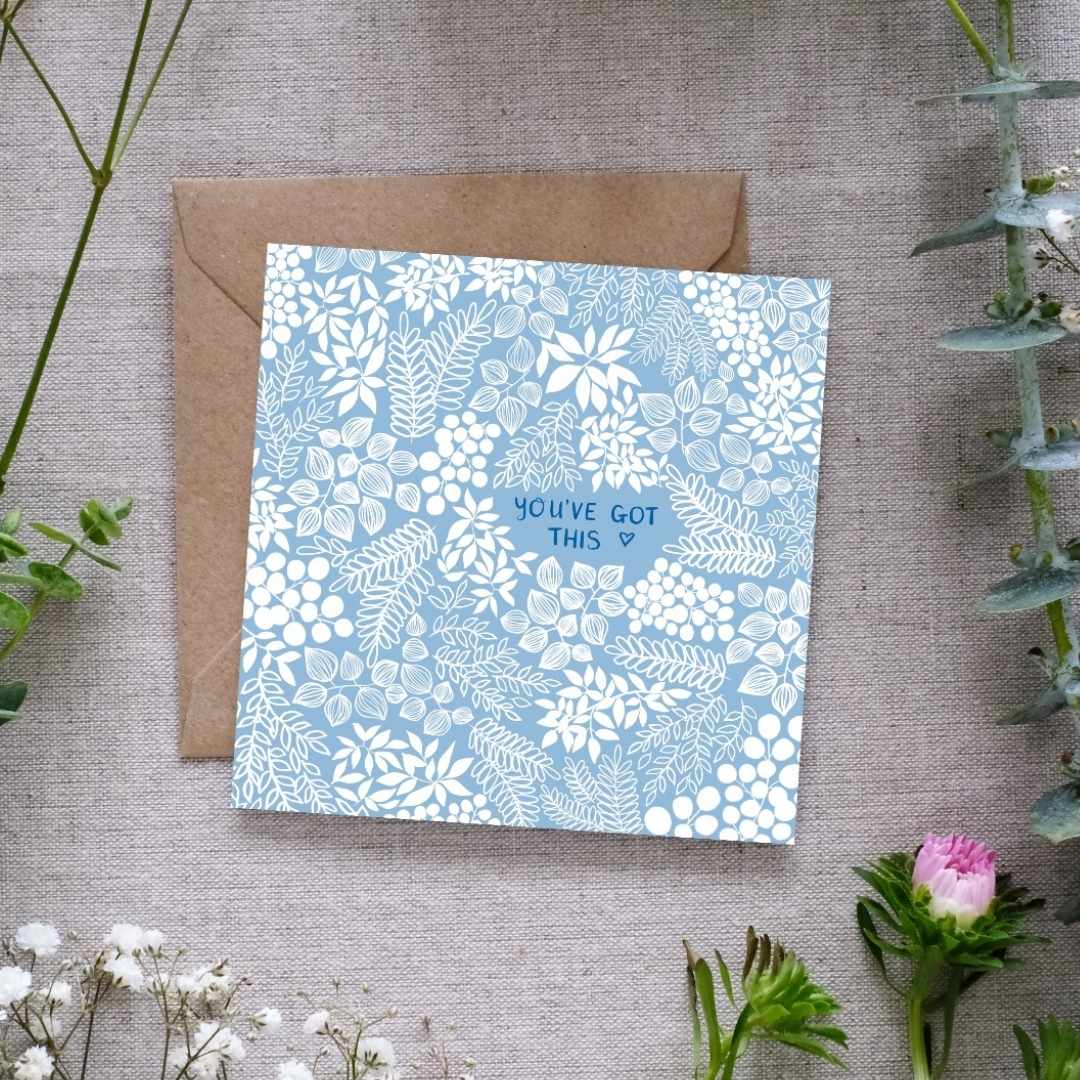 A square greeting card with a soft baby sky blue background and intricate white floral patterns. The words 'You've Got This' are centered in our hand-writing with a tiny heart outline beside it. The card is placed on a textured linen surface, partially resting on a brown kraft envelope. Surrounding the card are sprigs of eucalyptus, baby’s breath, and other greenery, along with a small pink flower.