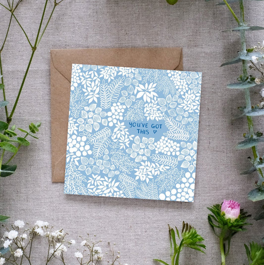 A square greeting card with a soft baby sky blue background and intricate white floral patterns. The words 'You've Got This' are centered in our hand-writing with a tiny heart outline beside it. The card is placed on a textured linen surface, partially resting on a brown kraft envelope. Surrounding the card are sprigs of eucalyptus, baby’s breath, and other greenery, along with a small pink flower.