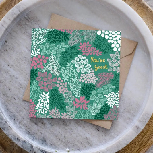 Image of a greeting card with a brown Kraft envelope behind it. It's set on a grey plate, with a wooden circular board underneath. There are sprigs of baby's breath and eucalyptus visible too. 
The greeting card is green with gold writing which says "You're Great". Surrounding the writing are clusters of leaves and flowers all in different shades of green, white and pink.