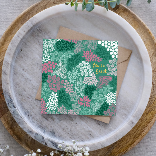 Image of a greeting card with a brown Kraft envelope behind it. It's set on a grey plate, with a wooden circular board underneath. There are sprigs of baby's breath and eucalyptus visible too. 
The greeting card is green with gold writing which says "You're Great". Surrounding the writing are clusters of leaves and flowers all in different shades of green, white and pink.