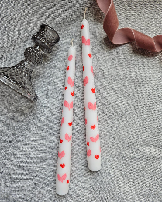 Two tapered dinner candles which are white with painted red hearts and pink leaves. They are lying on a grey cloth with a candle holder and a dusty pink ribbon beside them.