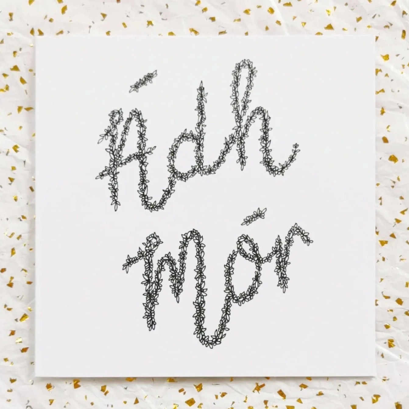 Image shows an Irish language greeting card that says Ádh Mór. The words ádh mór mean good luck in Irish and the words themselves are drawn entirely from a long string of leaves and petals. The illustration is done with black pen and the card itself is white. It is laid out on a background of white and gold paper for presentation.