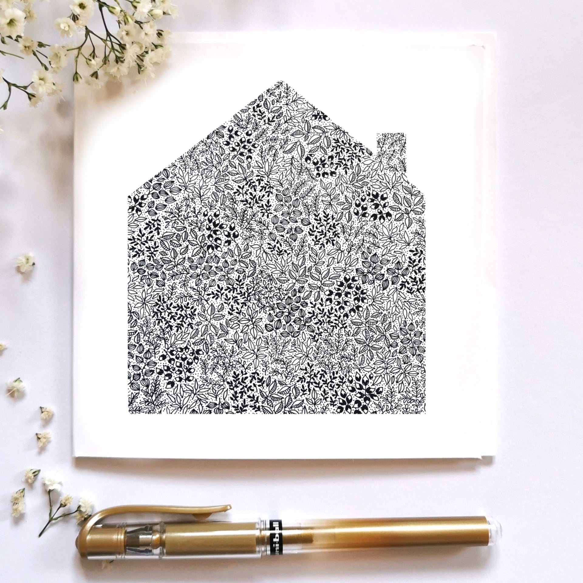 This is a picture of a black and white house made up of flowers and leaves. The house is intricately drawn with black pen so the details are small and delicate. The card is laid out beside some gypsophila flowers which are small, pretty and elegant, and beneath it lies a gold pen for presentation.