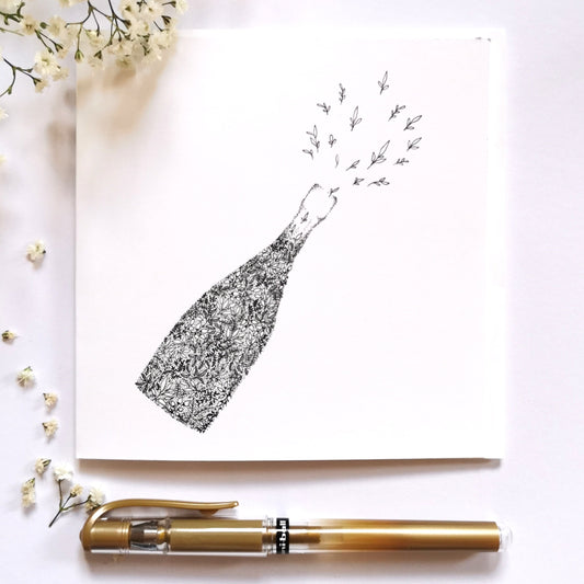 Image shows illustration of champagne bottle made entirely of black and white floral drawings. Bottle is slanted towards the right as if it had just been popped open with tiny flowers spritzing out the bottle as if it was the foam of a champagne bottle popping. Image is set on a cream table with a gold pen at the bottom of the image and white flowers on the left hand side of the image to show what illustration would look dressed up