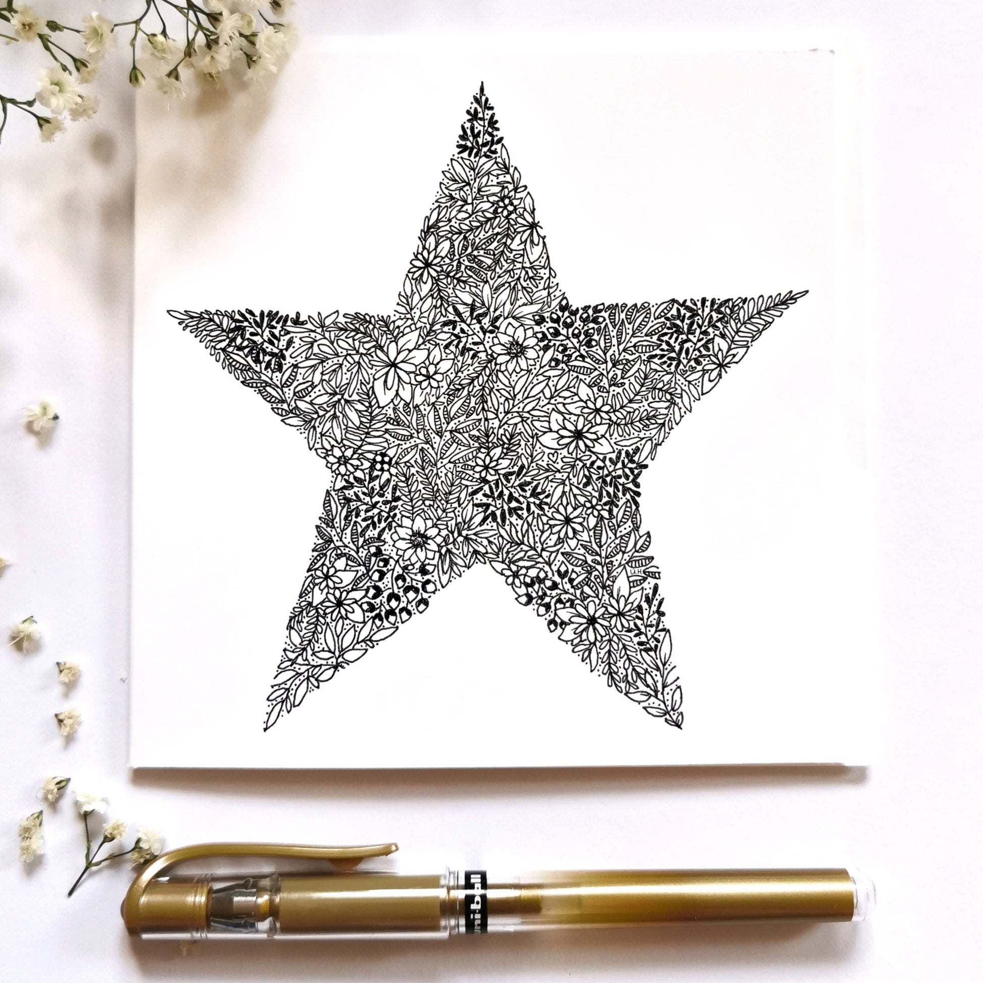 Image shows a star made from floral drawings. Image is entirely black and white and has dozens of different flowers leaves etc to make up this drawing. Image is laid on a cream surface with white flowers and gold pen to dress up image.